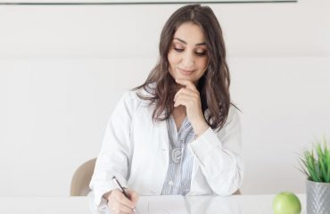 A female nutritionist in a lab coat writing notes with an apple and plant on the desk.